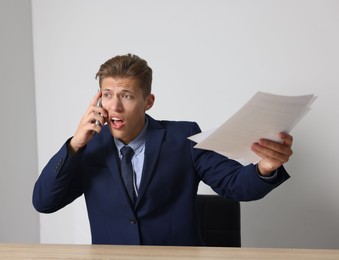 Photo of Young businessman with papers complaining to someone via phone at wooden table indoors
