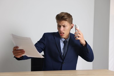 Photo of Young businessman with papers complaining to someone via phone at wooden table indoors