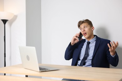 Photo of Young businessman with laptop complaining to someone via phone at wooden table indoors