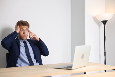 Photo of Young businessman with laptop complaining to someone via phone at wooden table indoors