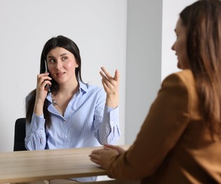 Photo of Young woman with phone complaining to businesswoman at wooden table indoors
