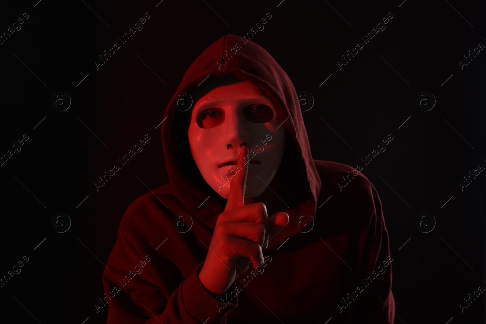 Photo of Anonymity. Masked man showing hush gesture on dark background with red light