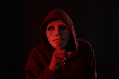 Photo of Anonymity. Masked man showing hush gesture on dark background with red light