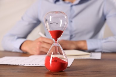 Photo of Deadline. Hourglass and man working at wooden table, selective focus