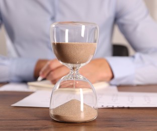 Photo of Deadline. Hourglass and man working at wooden table, selective focus