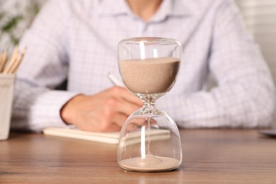 Photo of Deadline. Hourglass and man working at wooden table, selective focus