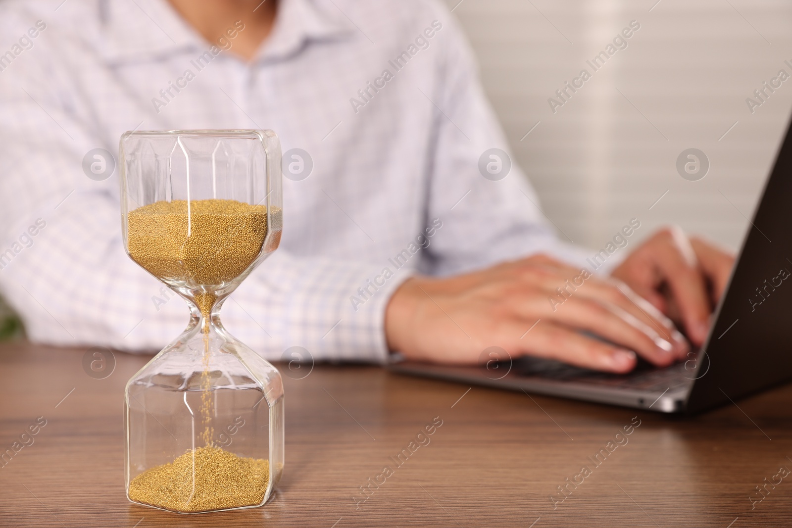 Photo of Deadline. Hourglass and man working with laptop at wooden table, selective focus