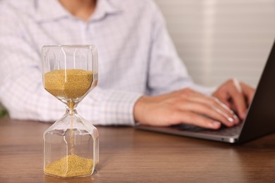 Photo of Deadline. Hourglass and man working with laptop at wooden table, selective focus