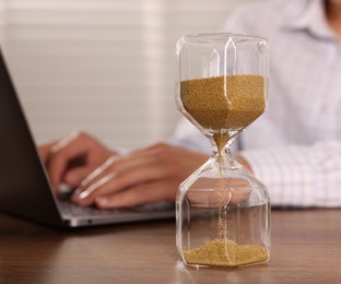 Photo of Deadline. Hourglass and man working with laptop at wooden table, selective focus