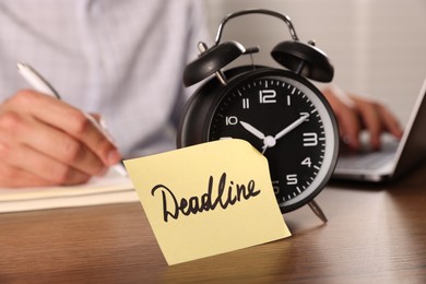 Photo of Paper note with word Deadline on alarm clock and man working at wooden table, selective focus