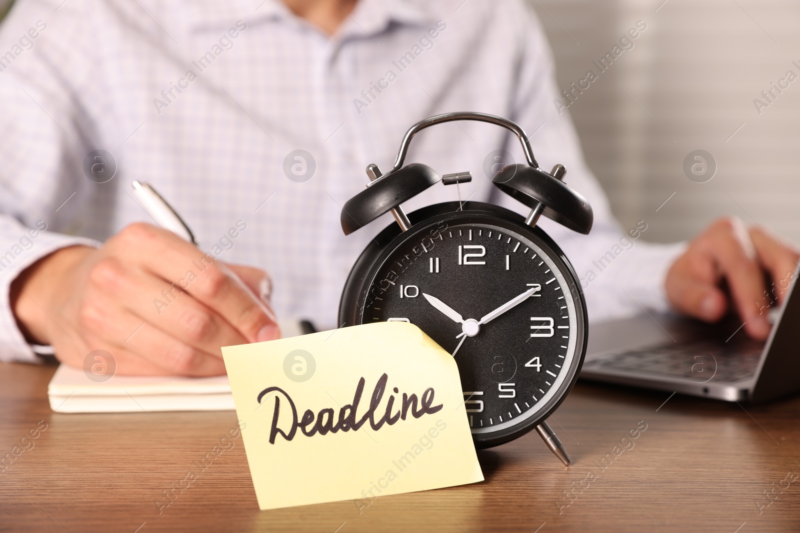 Photo of Paper note with word Deadline on alarm clock and man working at wooden table, selective focus