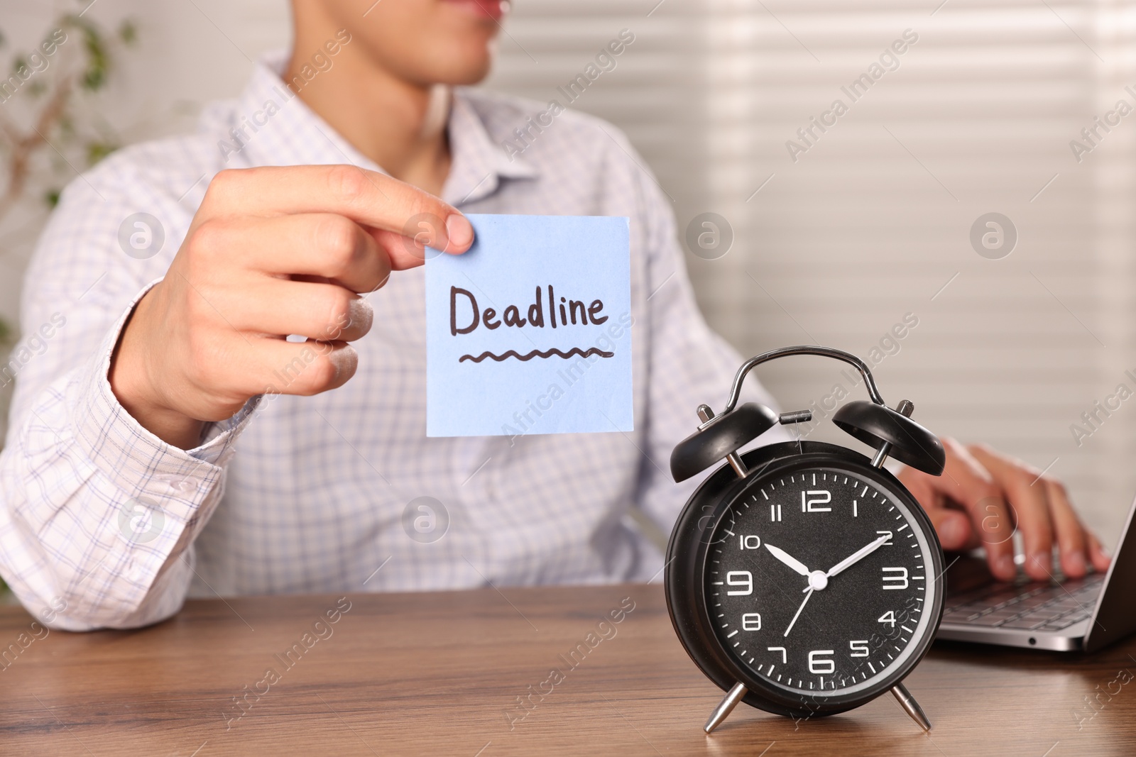 Photo of Man holding paper note with word Deadline while working and alarm clock on wooden table, selective focus