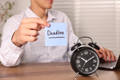 Photo of Man holding paper note with word Deadline while working and alarm clock on wooden table, selective focus
