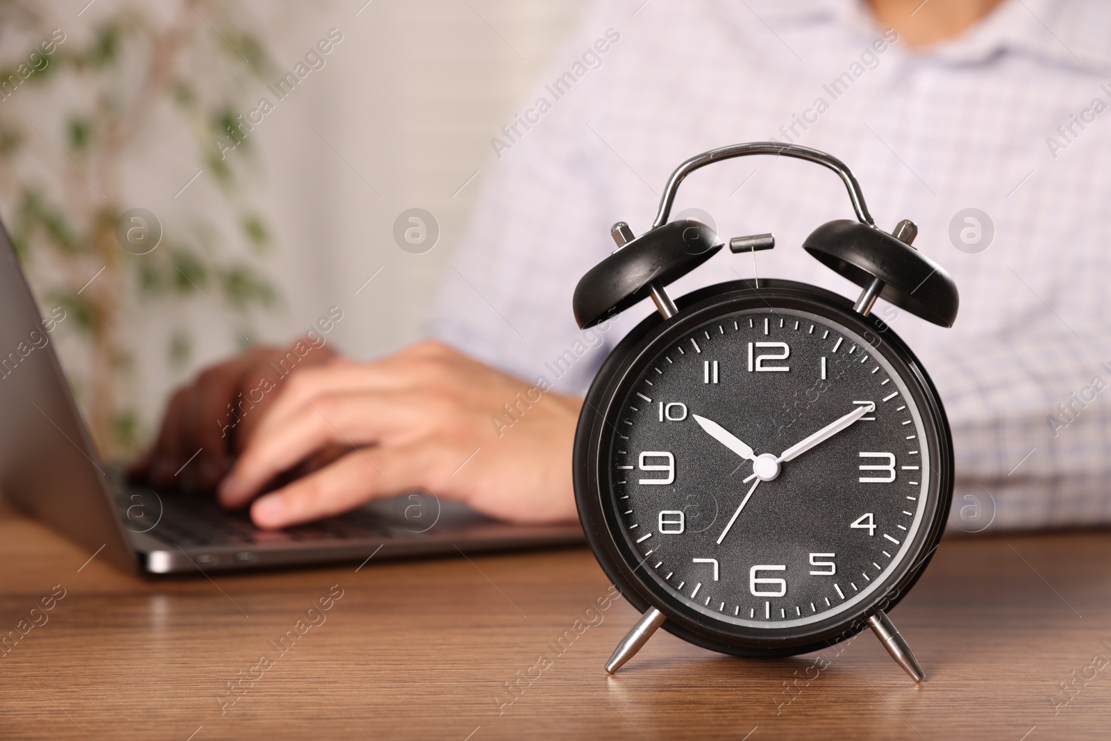 Deadline. Alarm clock and man working on laptop at wooden table, selective focus Photo of Deadline. Alarm clock and man working on laptop at wooden table, selective focus