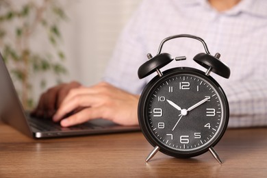 Photo of Deadline. Alarm clock and man working on laptop at wooden table, selective focus