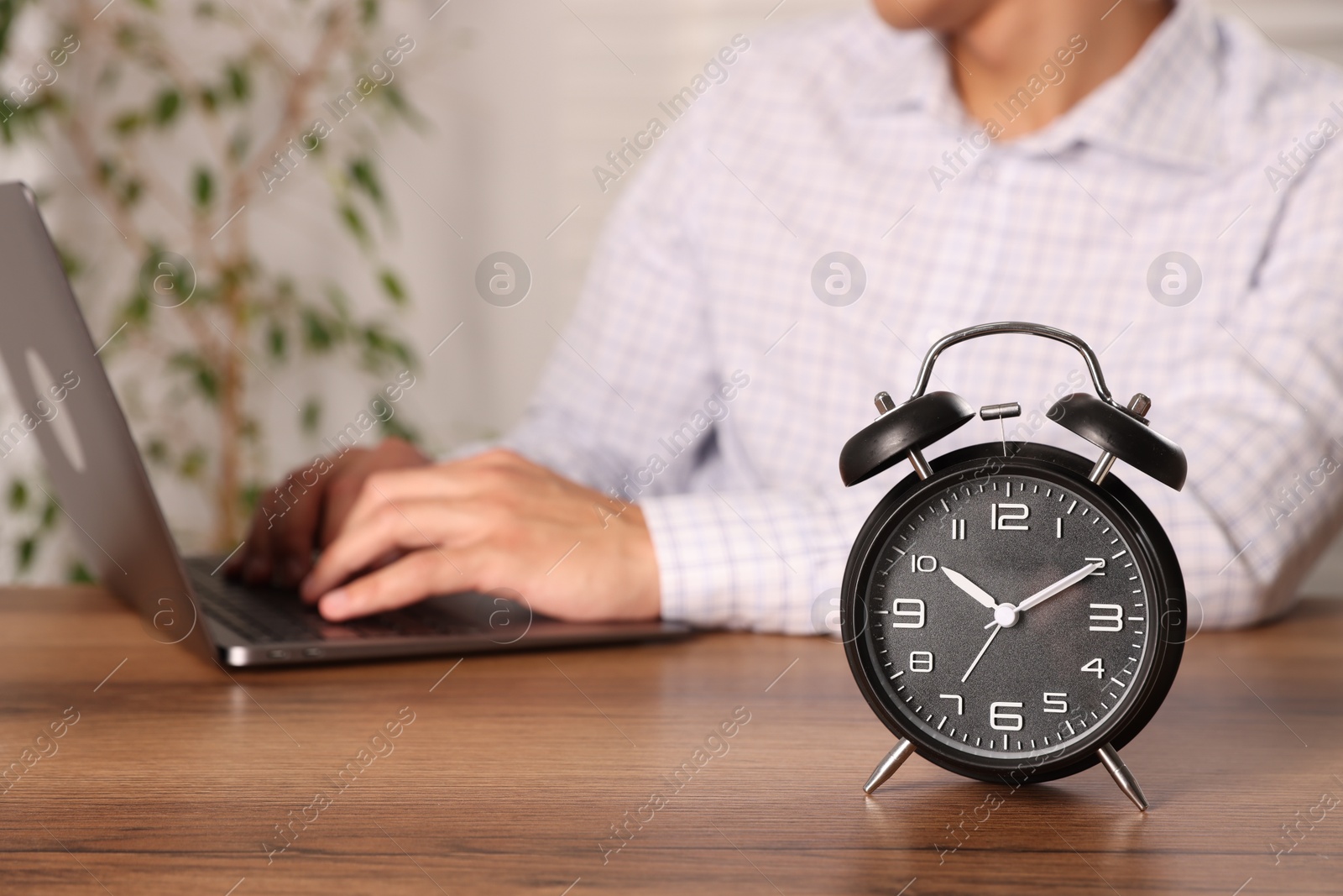 Photo of Deadline. Alarm clock and man working on laptop at wooden table, selective focus