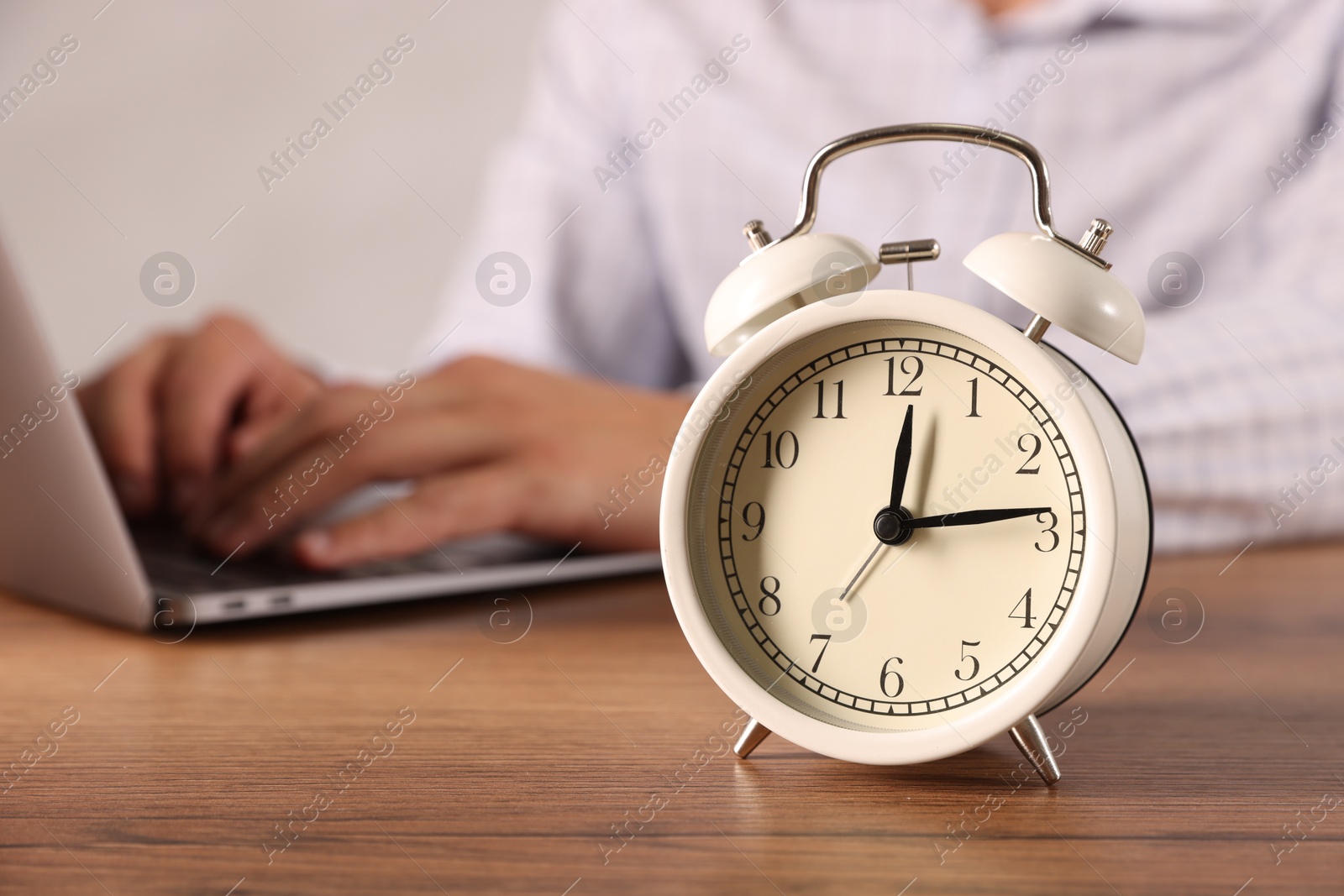 Photo of Deadline. Alarm clock and man working on laptop at wooden table, selective focus