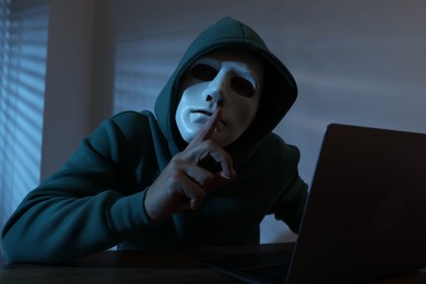 Photo of Anonymity. Masked man with laptop showing hush gesture at table in dark