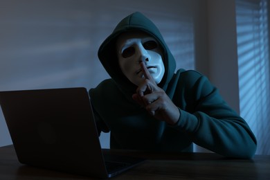 Photo of Anonymity. Masked man with laptop showing hush gesture at table in dark