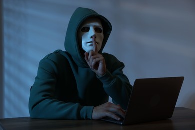 Photo of Anonymity. Masked man with laptop showing hush gesture at table in dark