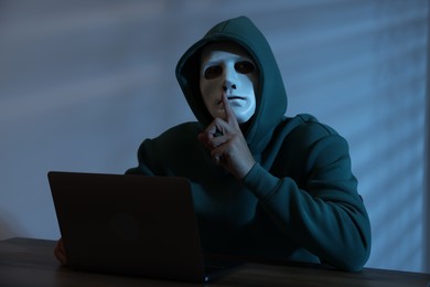 Photo of Anonymity. Masked man with laptop showing hush gesture at table in dark