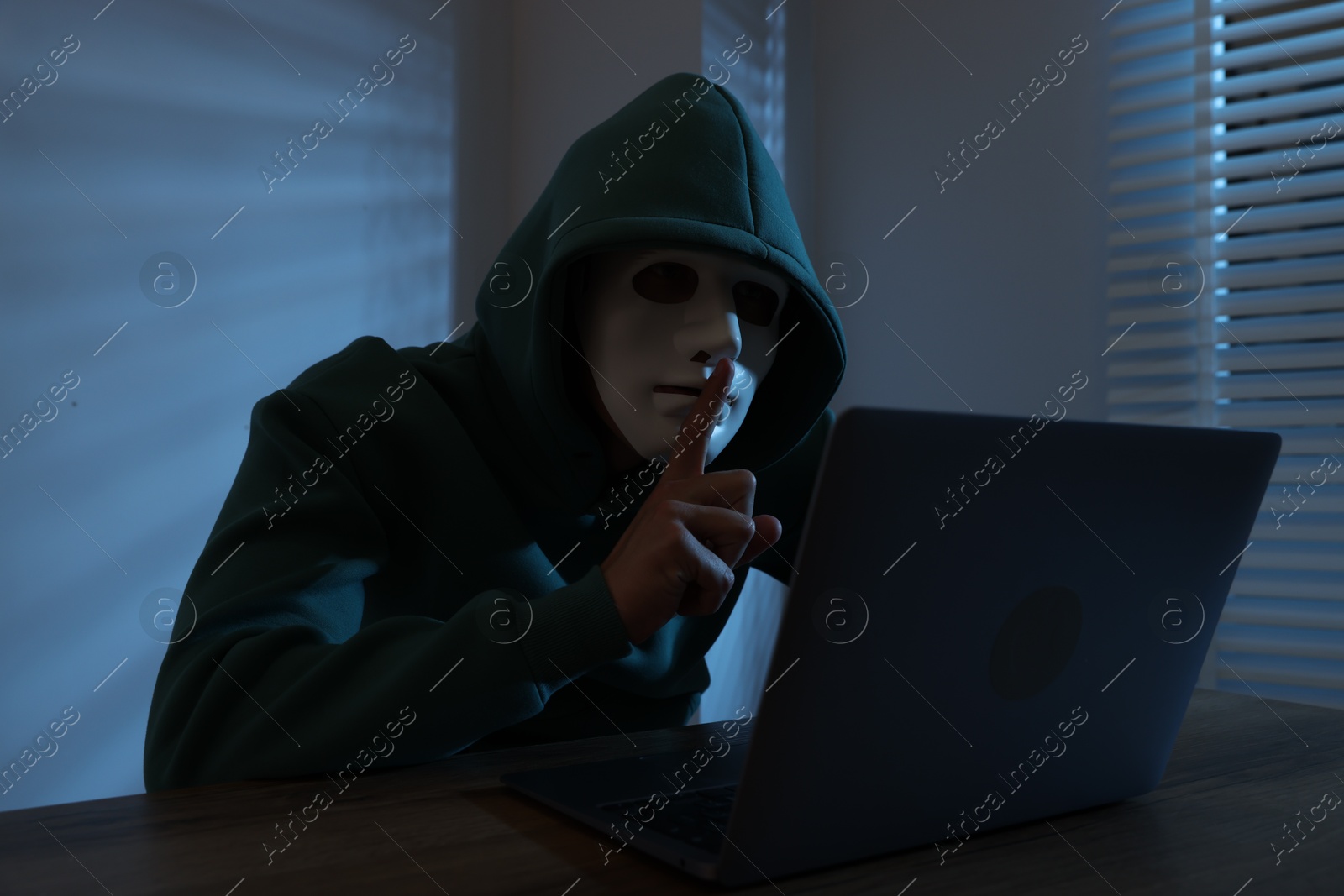 Photo of Anonymity. Masked man with laptop showing hush gesture at table in dark