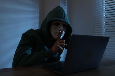 Photo of Anonymity. Masked man with laptop showing hush gesture at table in dark