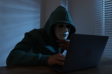 Photo of Anonymity. Masked man with laptop at table in dark