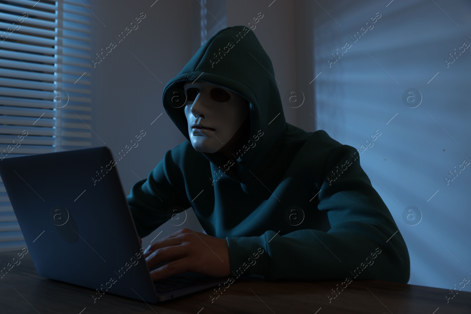 Photo of Anonymity. Masked man with laptop at table in dark