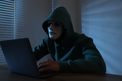 Photo of Anonymity. Masked man with laptop at table in dark