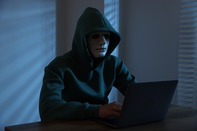 Photo of Anonymity. Masked man with laptop at table in dark