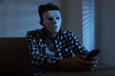Photo of Anonymity. Masked man with smartphone and laptop at table in dark