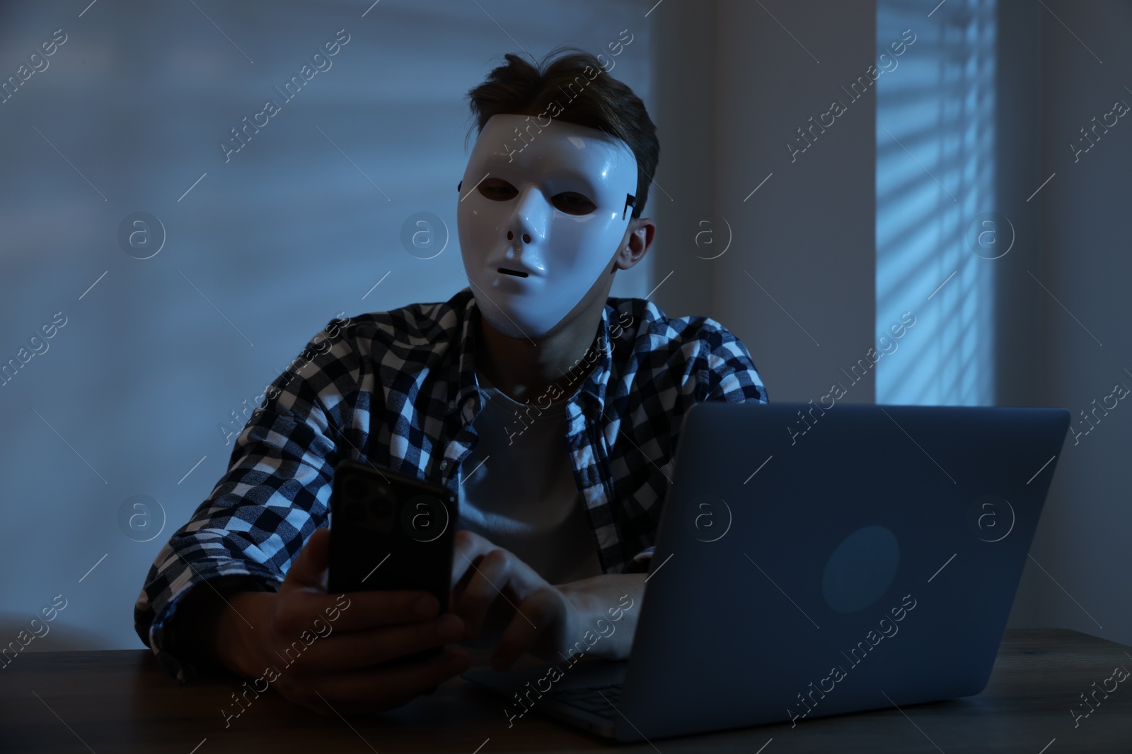Photo of Anonymity. Masked man with smartphone and laptop at table in dark