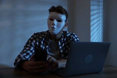 Photo of Anonymity. Masked man with smartphone and laptop at table in dark