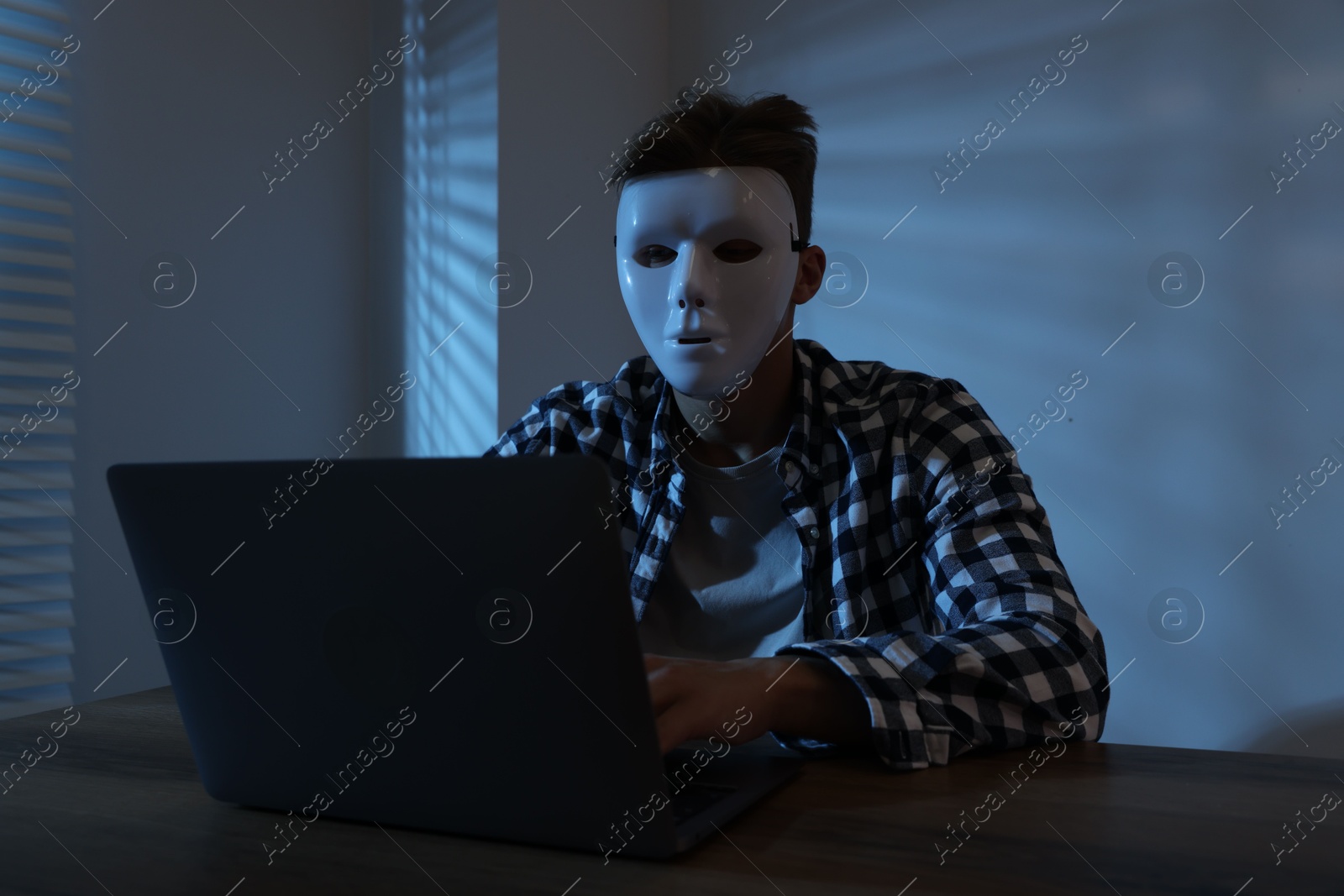 Photo of Anonymity. Masked man with laptop at table in dark
