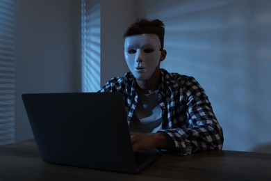 Photo of Anonymity. Masked man with laptop at table in dark