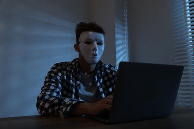 Photo of Anonymity. Masked man with laptop at table in dark