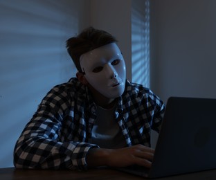 Photo of Anonymity. Masked man with laptop at table in dark