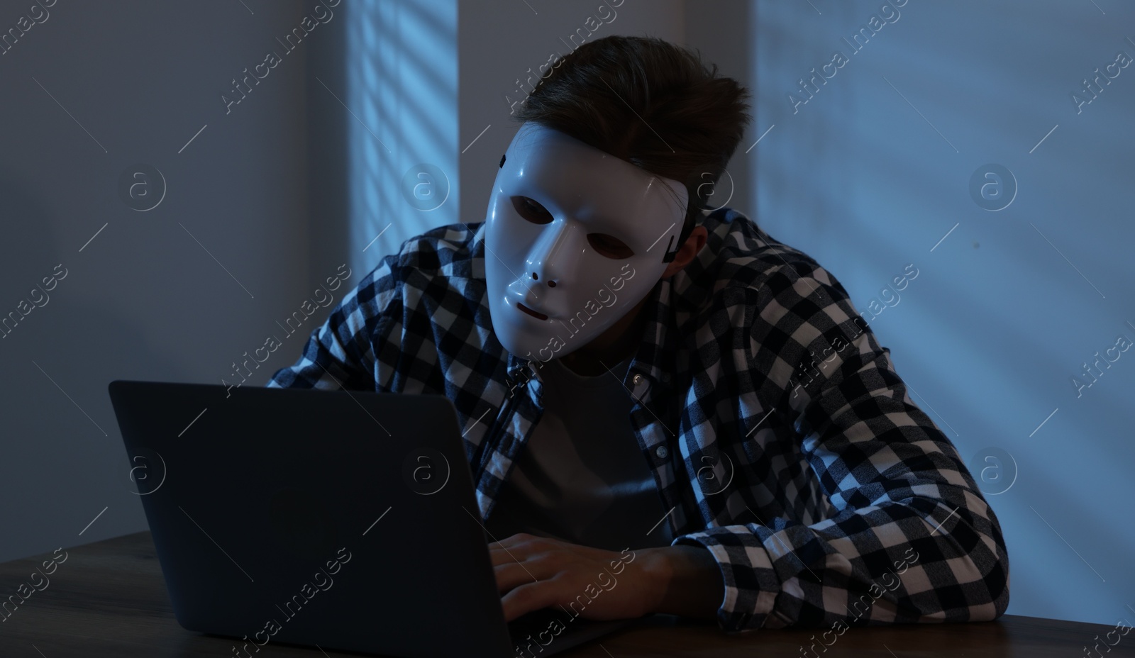 Photo of Anonymity. Masked man with laptop at table in dark