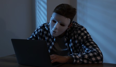 Photo of Anonymity. Masked man with laptop at table in dark