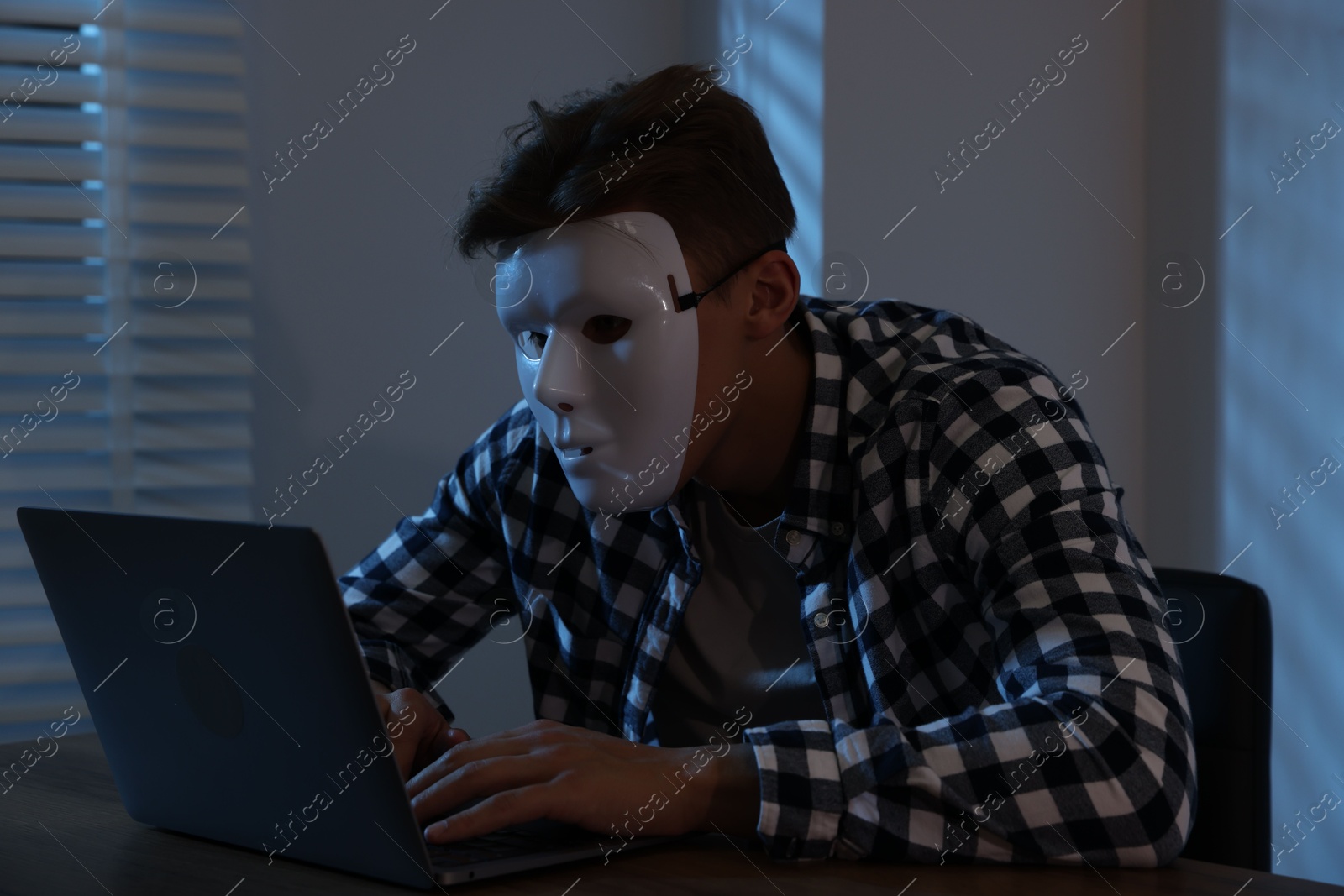 Photo of Anonymity. Masked man with laptop at table in dark