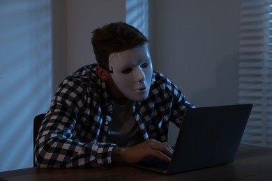 Photo of Anonymity. Masked man with laptop at table in dark