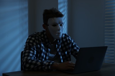 Photo of Anonymity. Masked man with laptop at table in dark