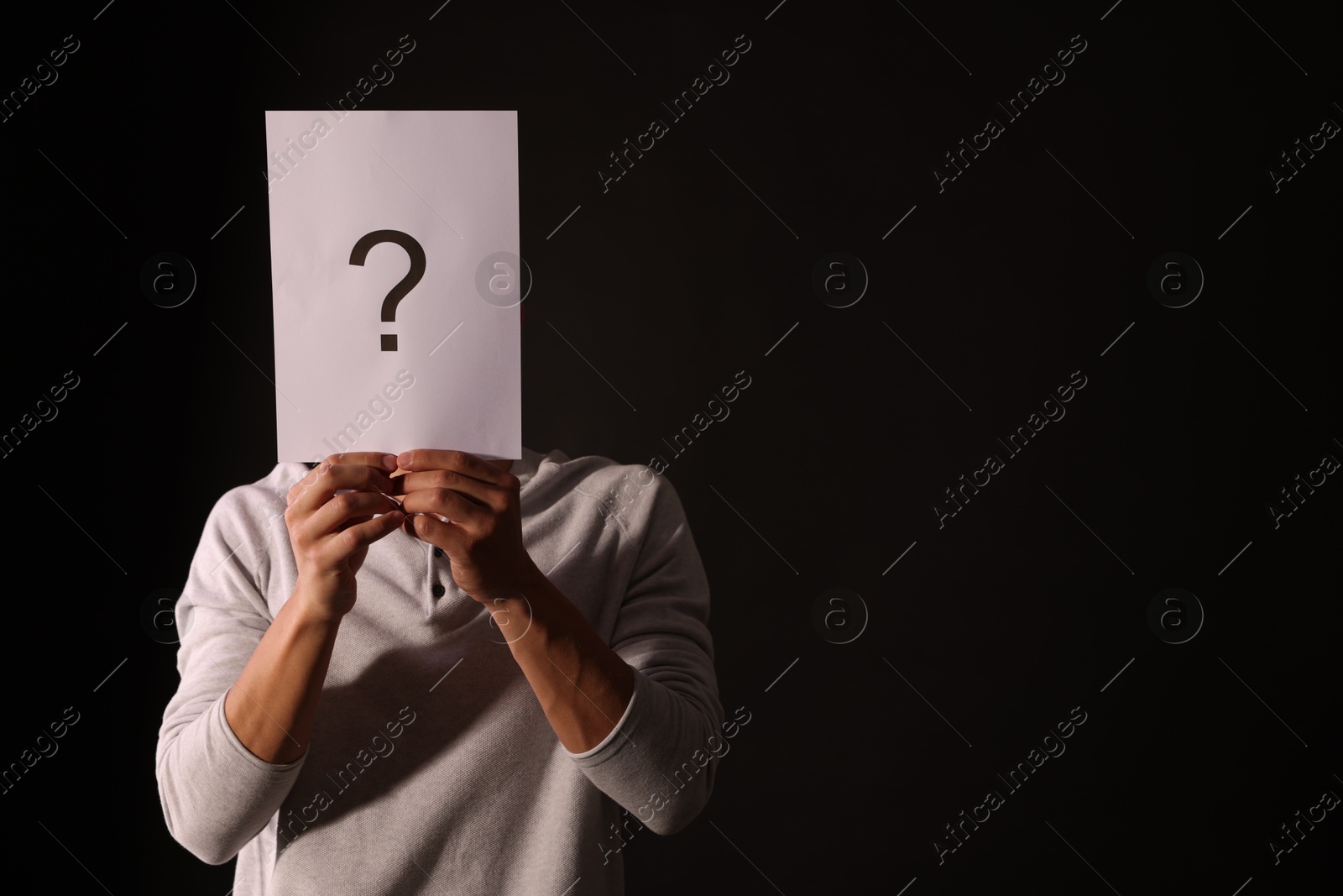 Photo of Man hiding behind paper sheet with question mark on dark background, space for text