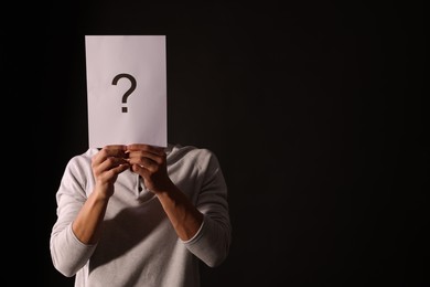 Photo of Man hiding behind paper sheet with question mark on dark background, space for text
