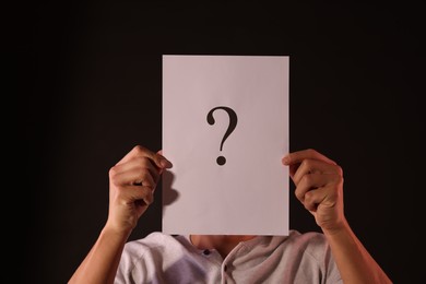 Photo of Man hiding behind paper sheet with question mark on dark background