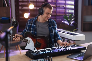 Photo of Smiling man in headphones working with MIDI keyboard and guitar at table in home studio