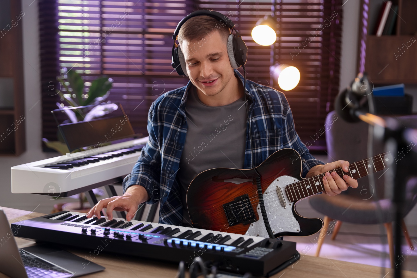 Photo of Smiling man in headphones working with MIDI keyboard and guitar at table in home studio