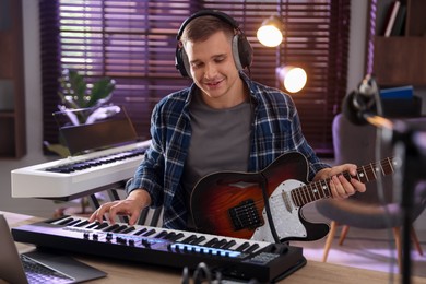Photo of Smiling man in headphones working with MIDI keyboard and guitar at table in home studio