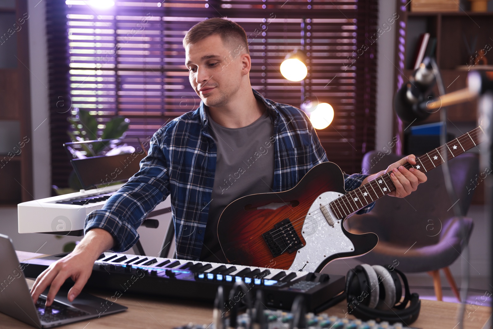 Photo of Man working with MIDI keyboard, laptop and guitar at table in home studio